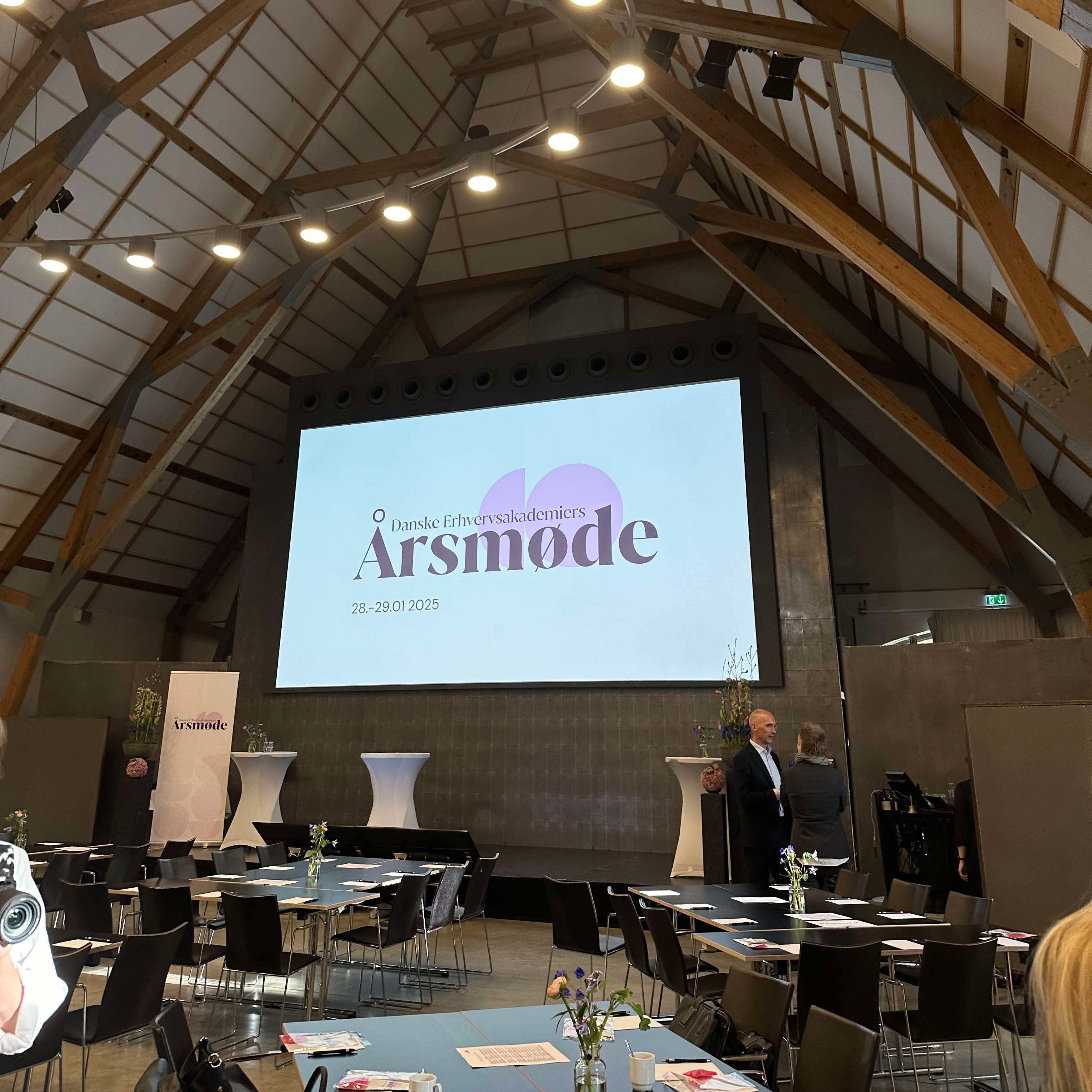 Three young professionals in a design meeting at a modern, open-concept workspace. They are seated around a round table with laptops, smartphones, and coffee cups, engaged in conversation and reviewing material samples. The setting includes glass walls, natural light, and a contemporary interior—suggesting a creative or academic environment, ideal for UX workshops or collaborative product design sessions.